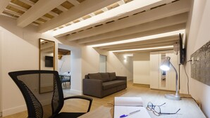A modern loft apartment with exposed wooden beams, a gray sofa, a black office chair, a desk with a notebook and glasses, and a large mirror. - Estudio Amor BRUJO by Cadiz4Rentals (Cádiz)