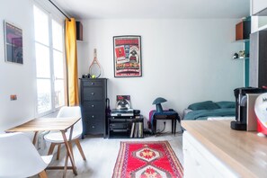 A compact living space with a bed, a desk, a chair, a small table, a record player, and a poster on the wall.
