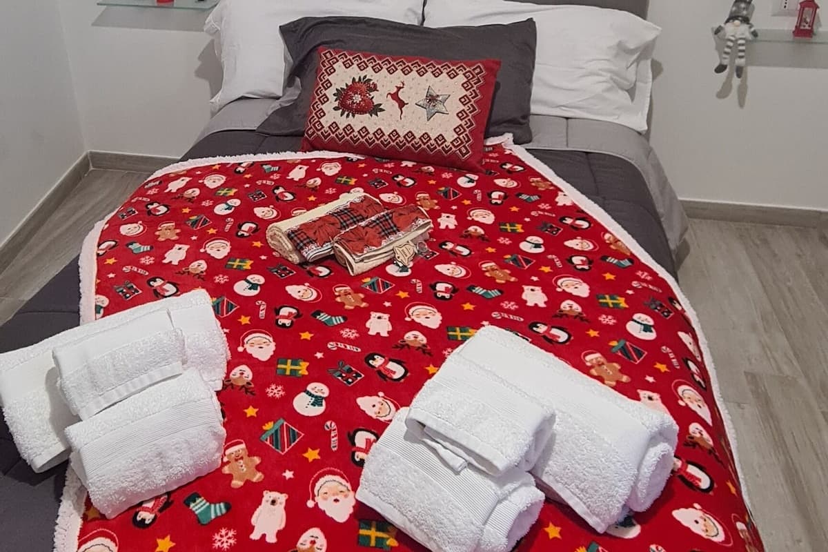 A neatly made bed with a red blanket featuring holiday-themed prints, white pillows, and two white towels.