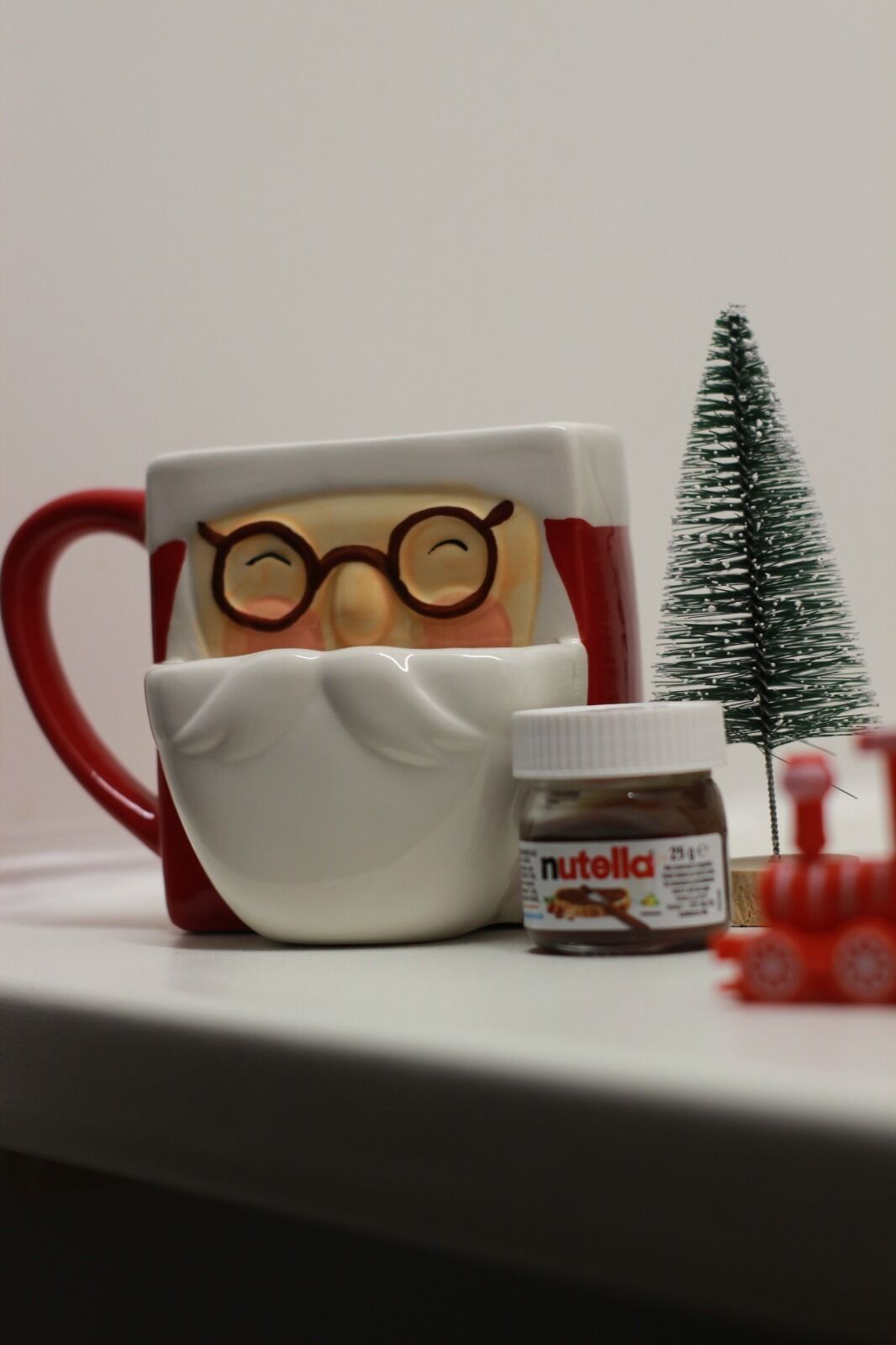 Een mok met een kerstman ontwerp, een pot Nutella en een kleine kerstboom op een tafel.
