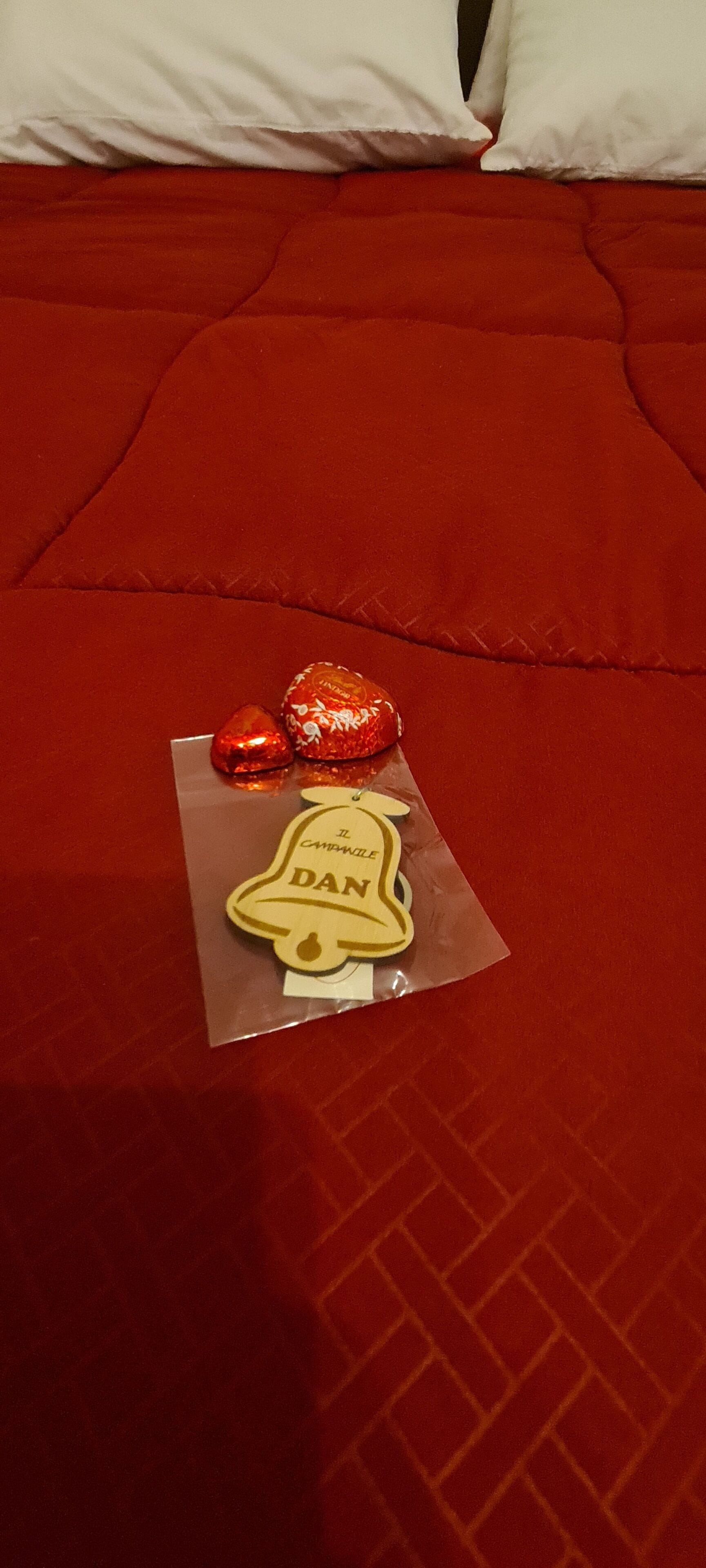 A bed with a red blanket, two wrapped chocolates, and a bell-shaped tag on a clear plastic sheet.