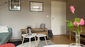 A cozy living room with a sofa, a coffee table, a small side table, and a door. There are framed pictures on the wall and a vase with a pink flower on a side table. - The Adelaide Boudoir, Charmant gîte avec lit king size et accès PMR (Vendresse)
