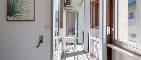Un balcón estrecho con una mesa y una silla blancos, un aire acondicionado portátil colgando, y vistas del edificio vecino.