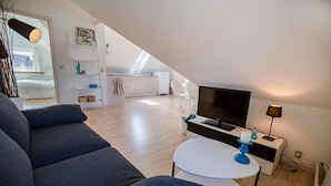 En modern lägenhet med ett vardagsrum som inrymmer en soffa, en TV och ett litet bord. Det finns ett köksområde med vita skåp och ett matbord. Lägenheten har ett sluttande tak och stora fönster.