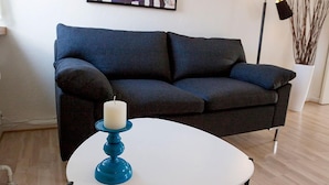 Ett modernt vardagsrum med en mörkgrå soffa, ett vitt runt bord med en blå ljusstake, en golvlampa och en inramad tecknad bild på väggen.