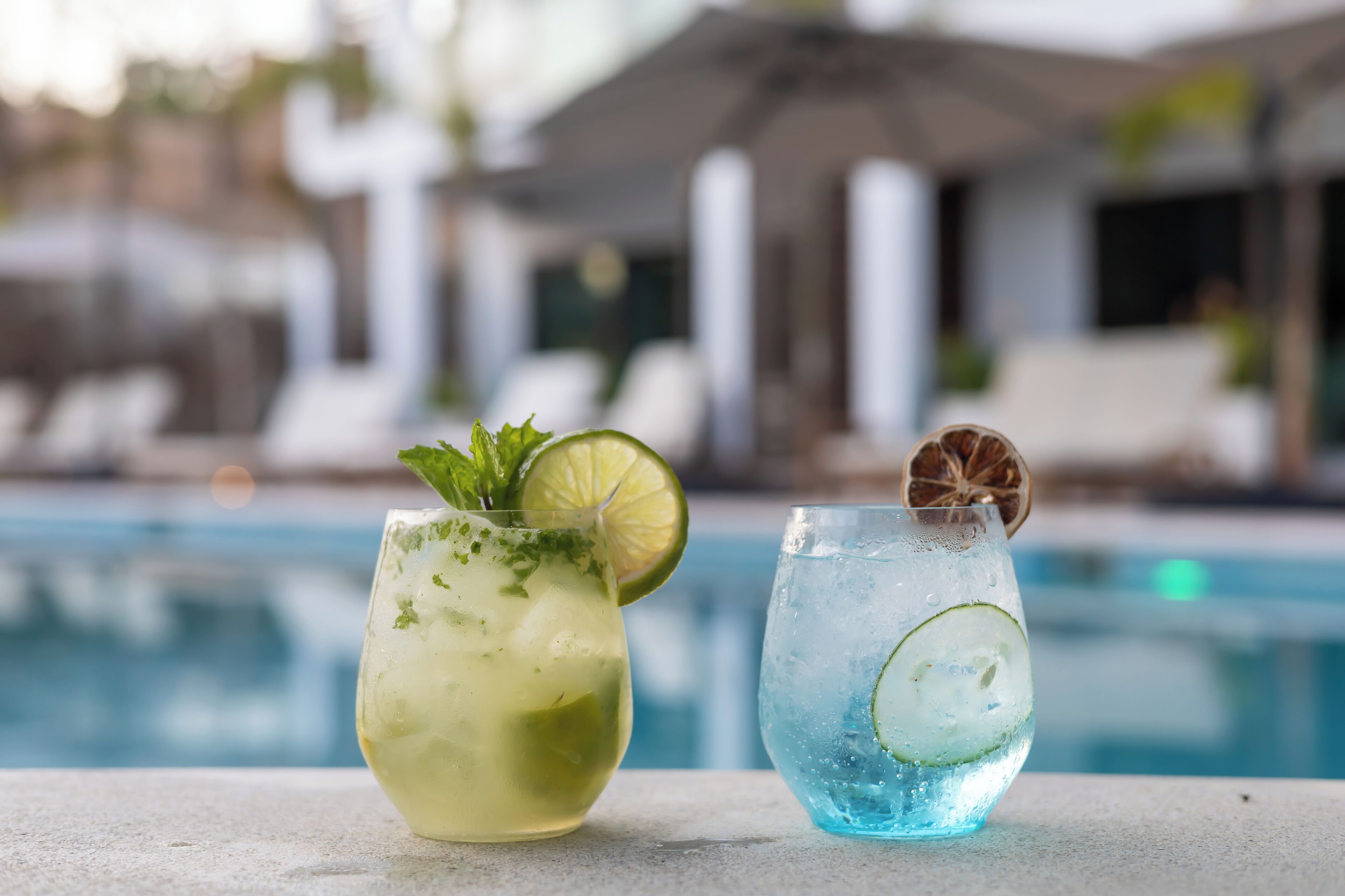 Dois copos de bebidas geladas com rodelas de limão e folhas de hortelã, colocados em uma mesa à beira da piscina, com piscina e cadeiras de sol ao fundo.