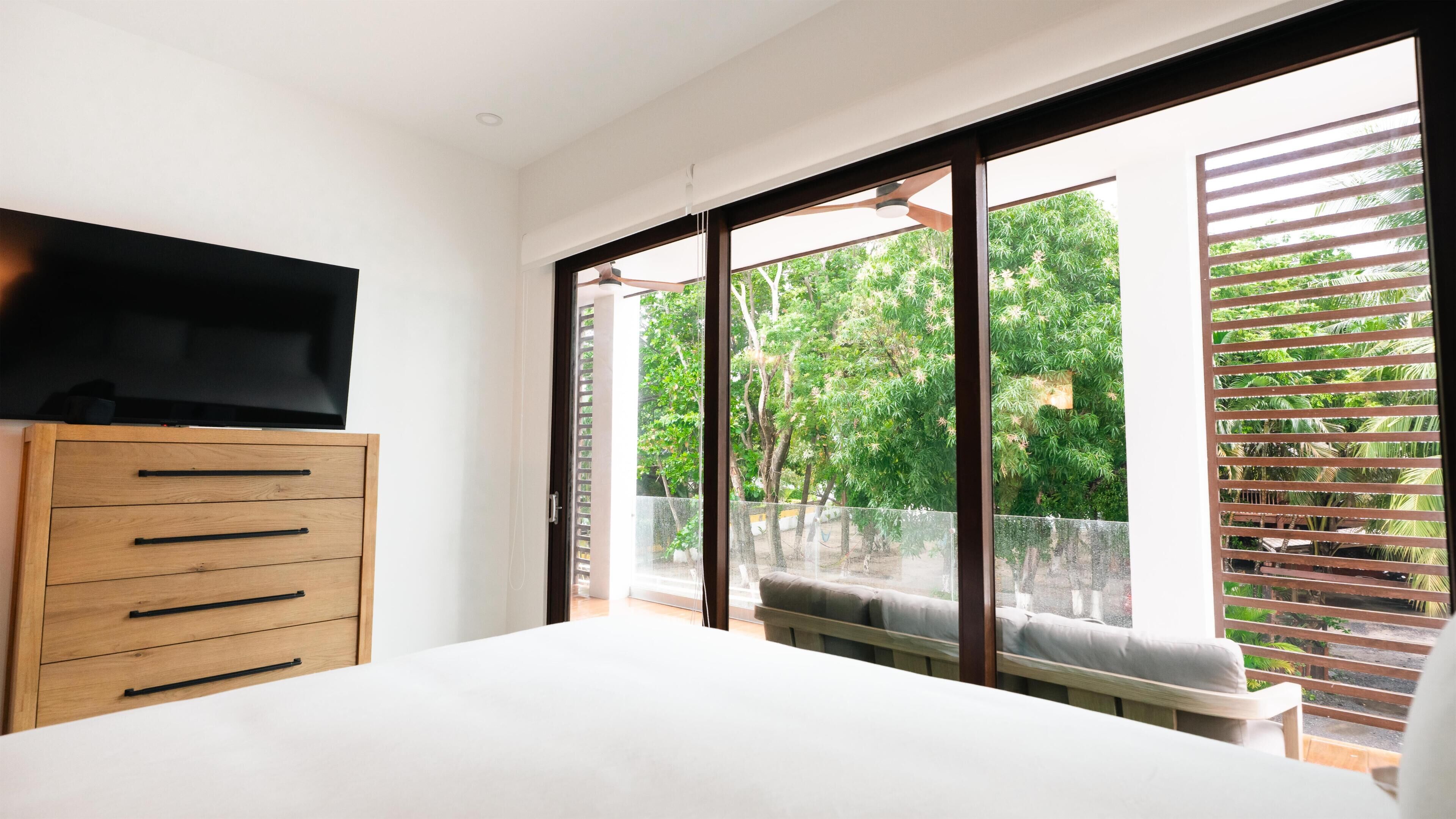 Un dormitorio con cama, cómoda de madera, televisor y balcón con vista a árboles.