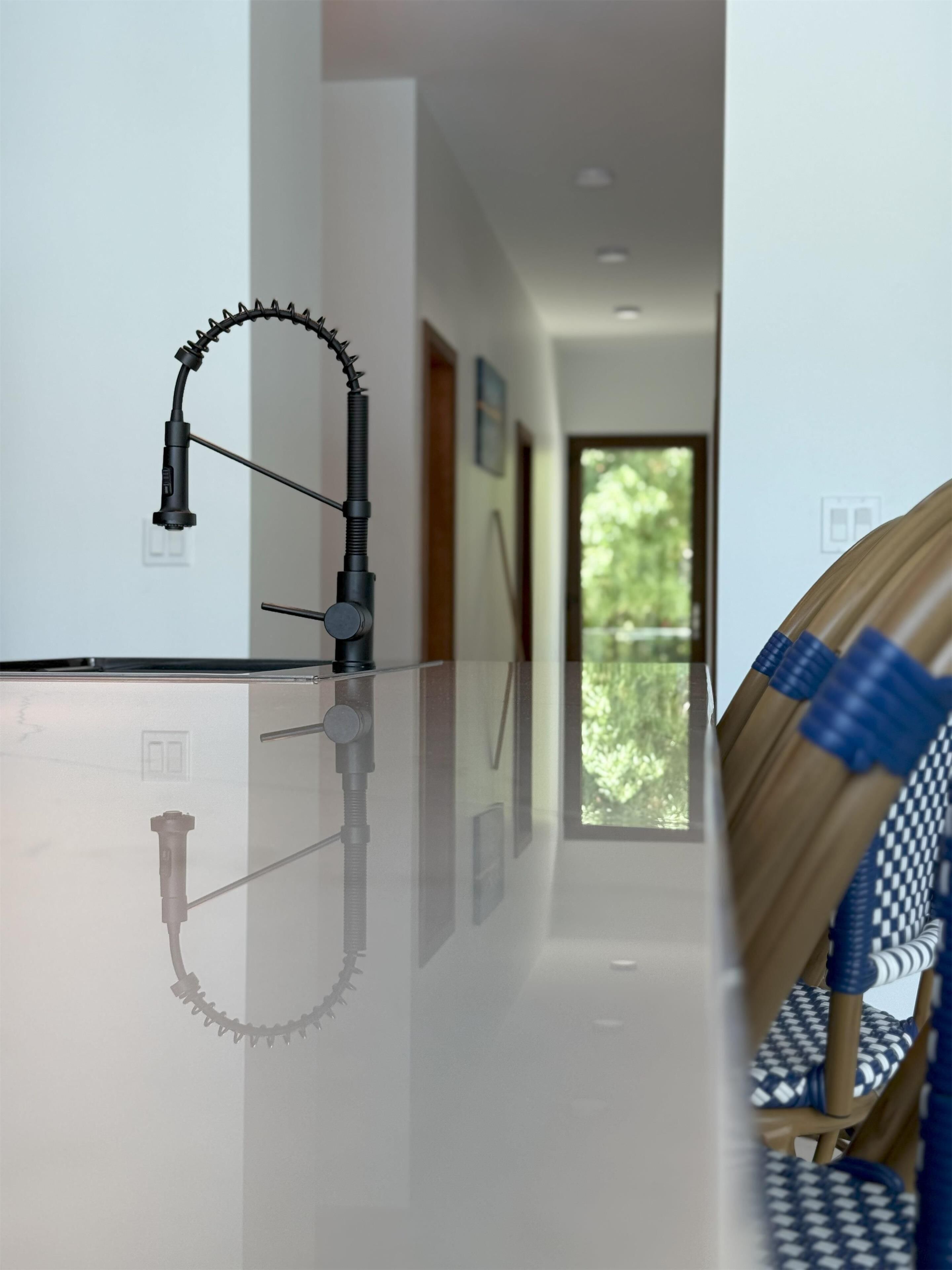 A modern kitchen with a sleek faucet, a reflective countertop, and a view of a wooden chair and a patterned cushion.