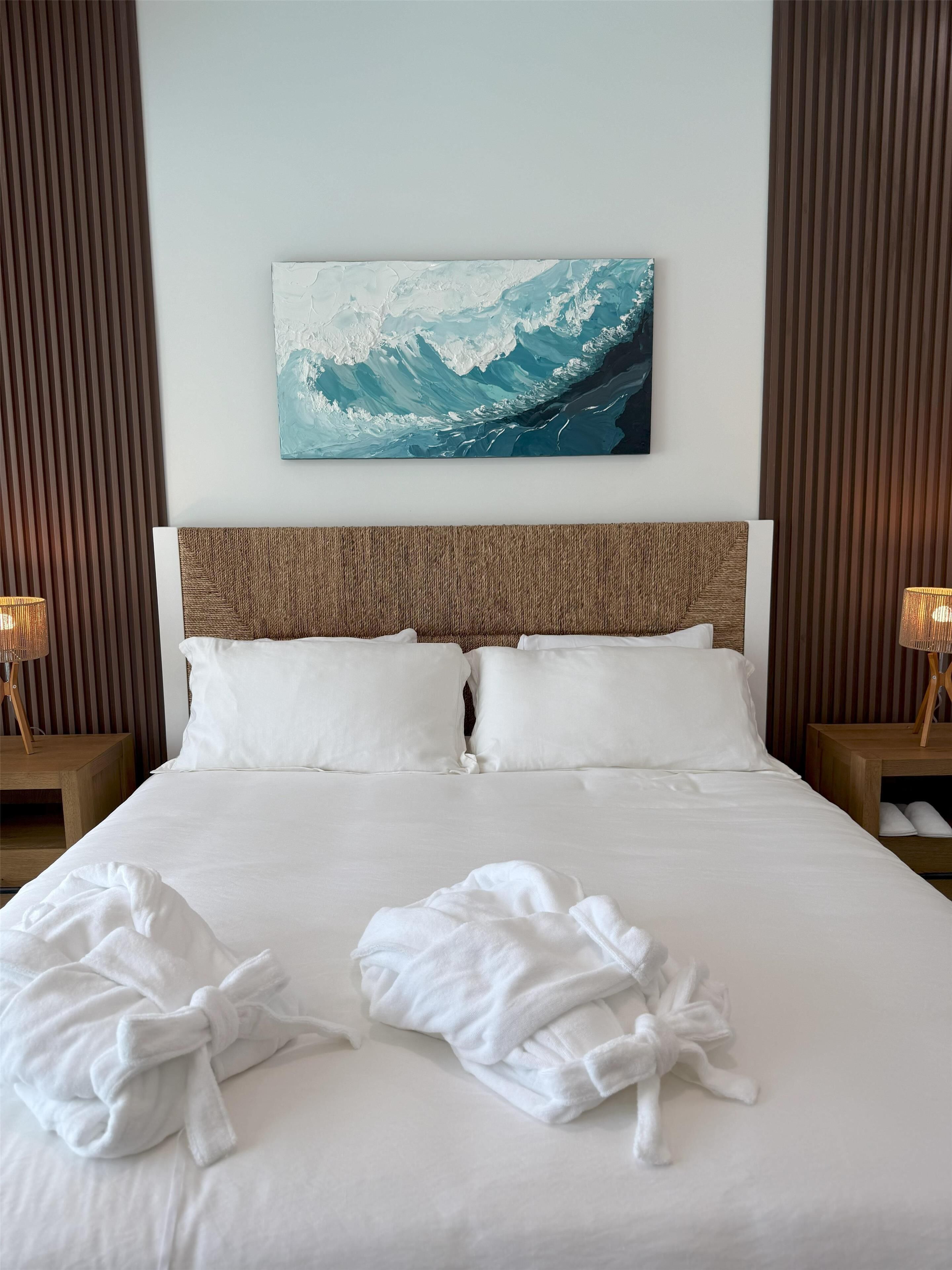 A hotel room with a large bed, two bedside tables with lamps, a painting of a wave on the wall, and a neatly made bed with two white bathrobes.