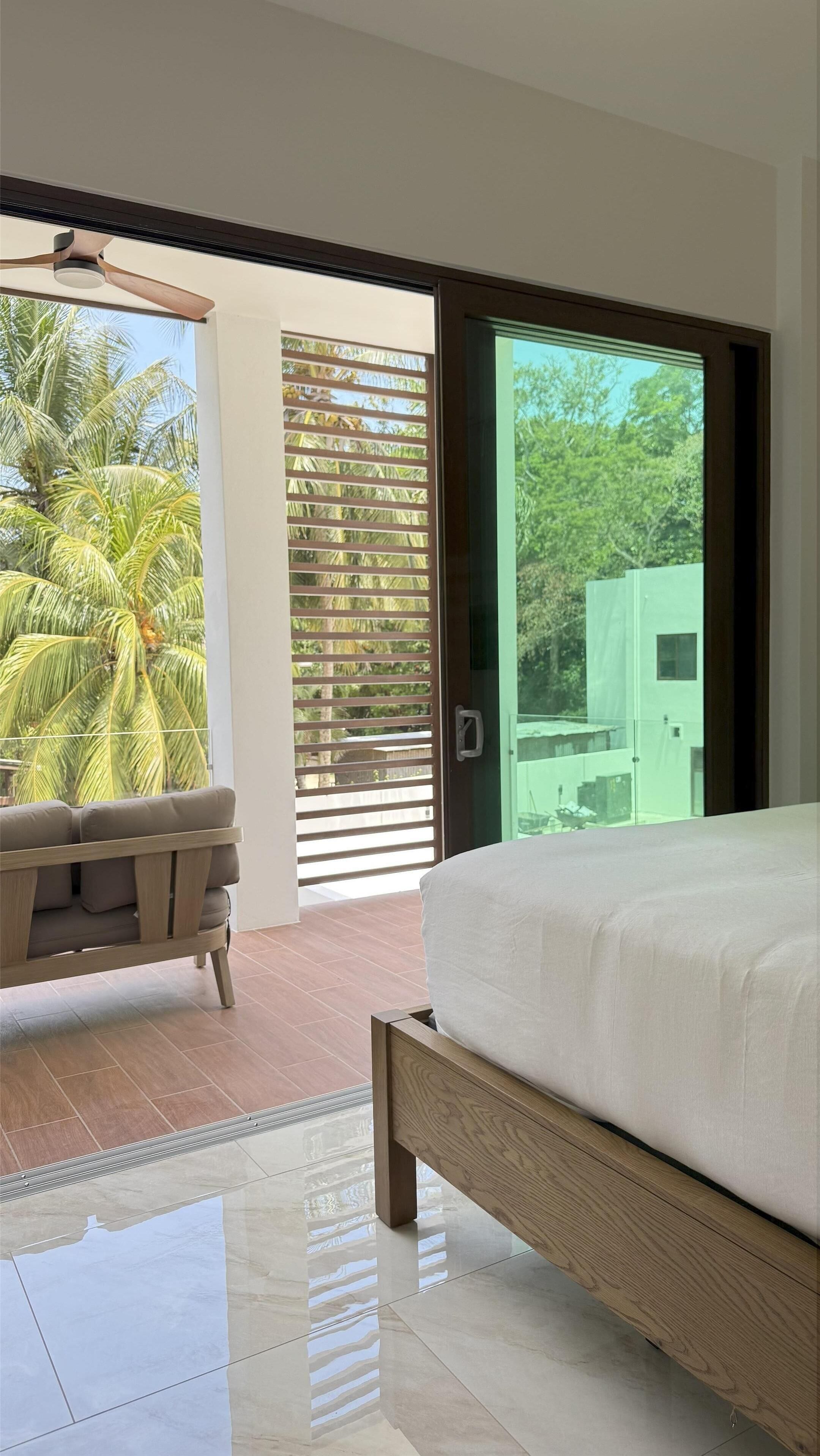 A bedroom with a bed, a sofa, a sliding glass door leading to a balcony with palm trees.