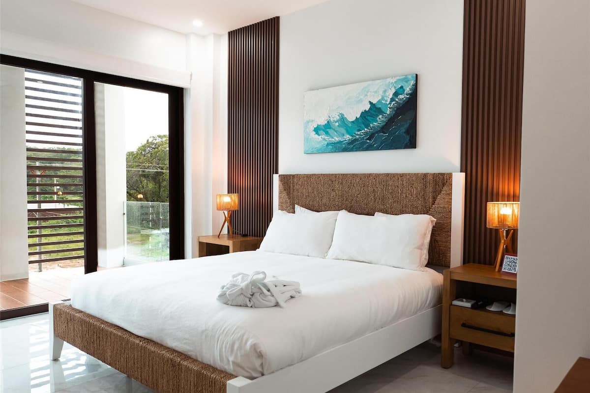 A modern bedroom with a large bed, a ceiling fan, a view through the glass door, and a painting on the wall.