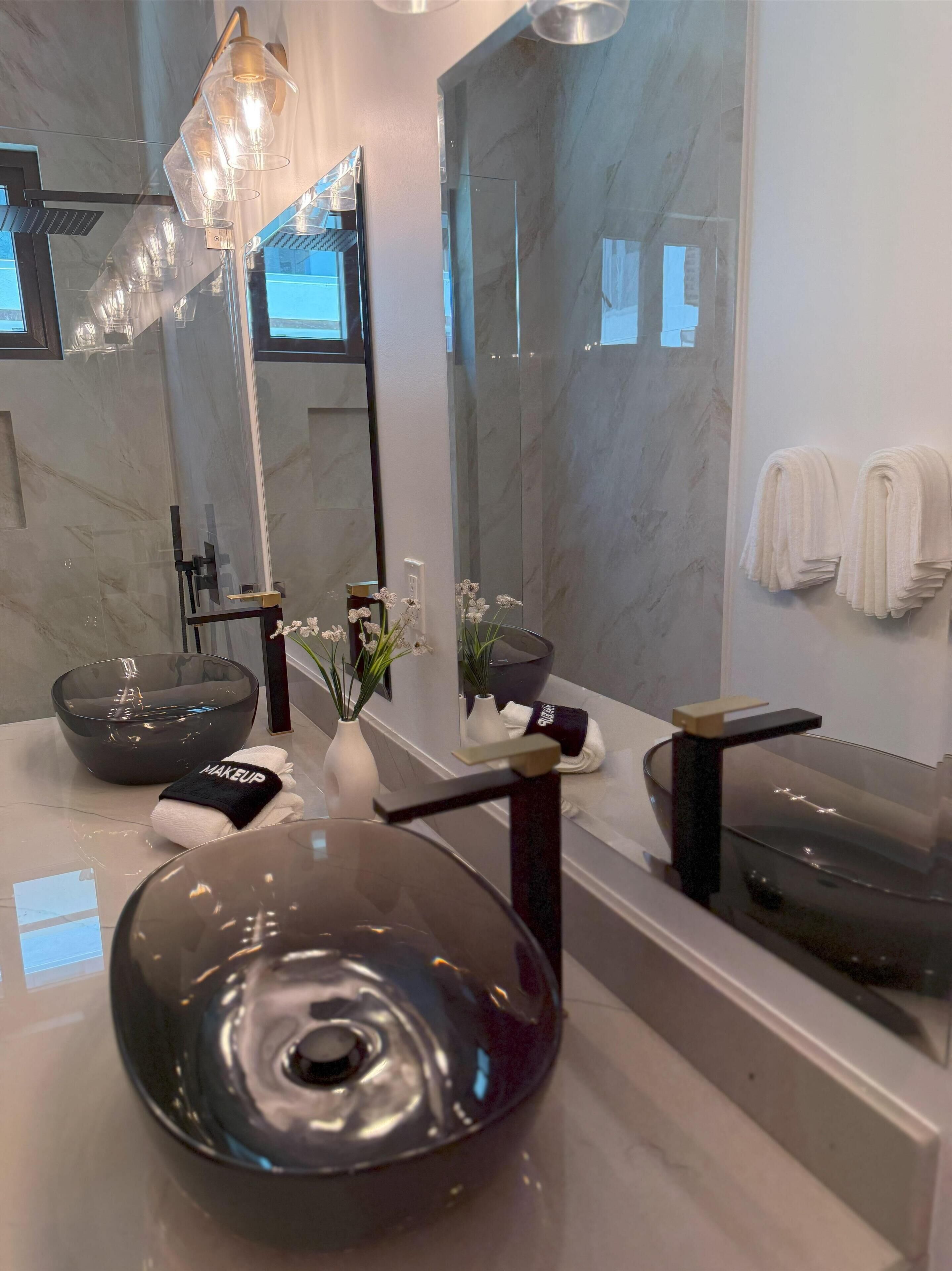 A modern bathroom with a large mirror, two bowls sink, a vase with flowers, and towels on the wall.