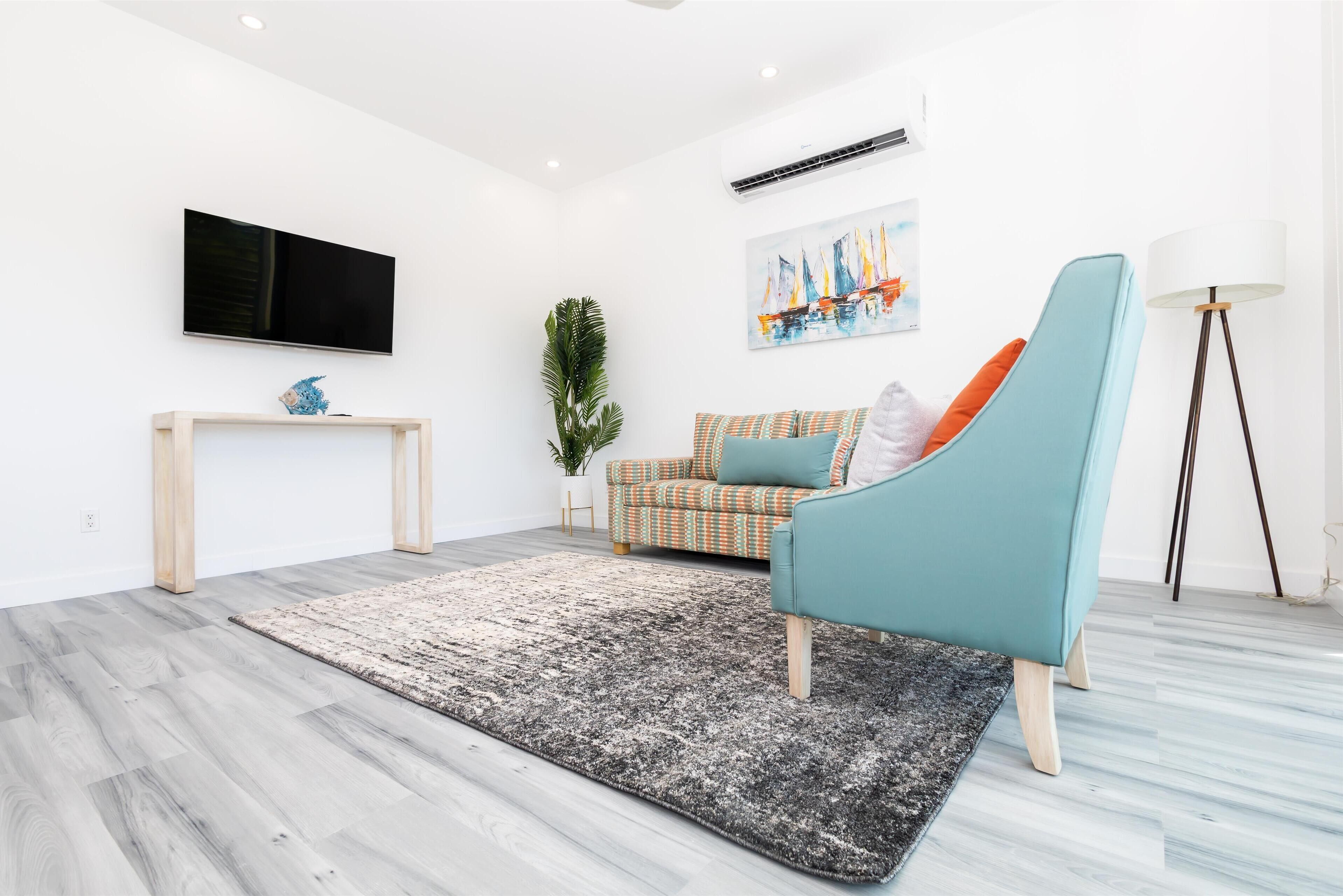 A modern living room with a flat-screen TV, a patterned sofa, a teal armchair, a floor lamp, and a large artwork on the wall.