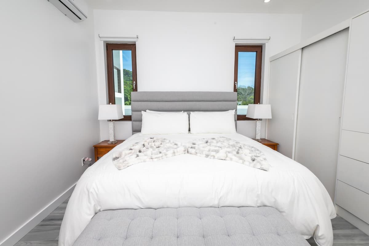 A bedroom with a bed, two bedside tables, two lamps, a window, and a view of greenery.