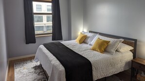 Een slaapkamer met een bed, een raam met gordijnen, een houten vloer en een vloerkleed.