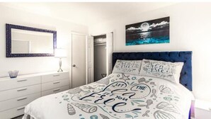 A bedroom with a bed featuring a white and blue patterned bedspread, a dark blue upholstered headboard, and a wall-mounted mirror. - Cozy beach cottage steps away from Hollywood Beach (Hollywood)