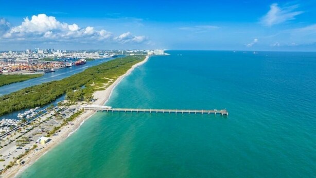 An aerial view of a coastal area with a long pier extending into the ocean, a beach, and a cityscape in the background. - Cozy beach cottage steps away from Hollywood Beach (Hollywood)