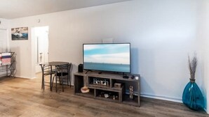 A living room with a flat-screen TV on a wooden stand, a dining table with chairs, and a vase with branches. - Cozy beach cottage steps away from Hollywood Beach (Hollywood)