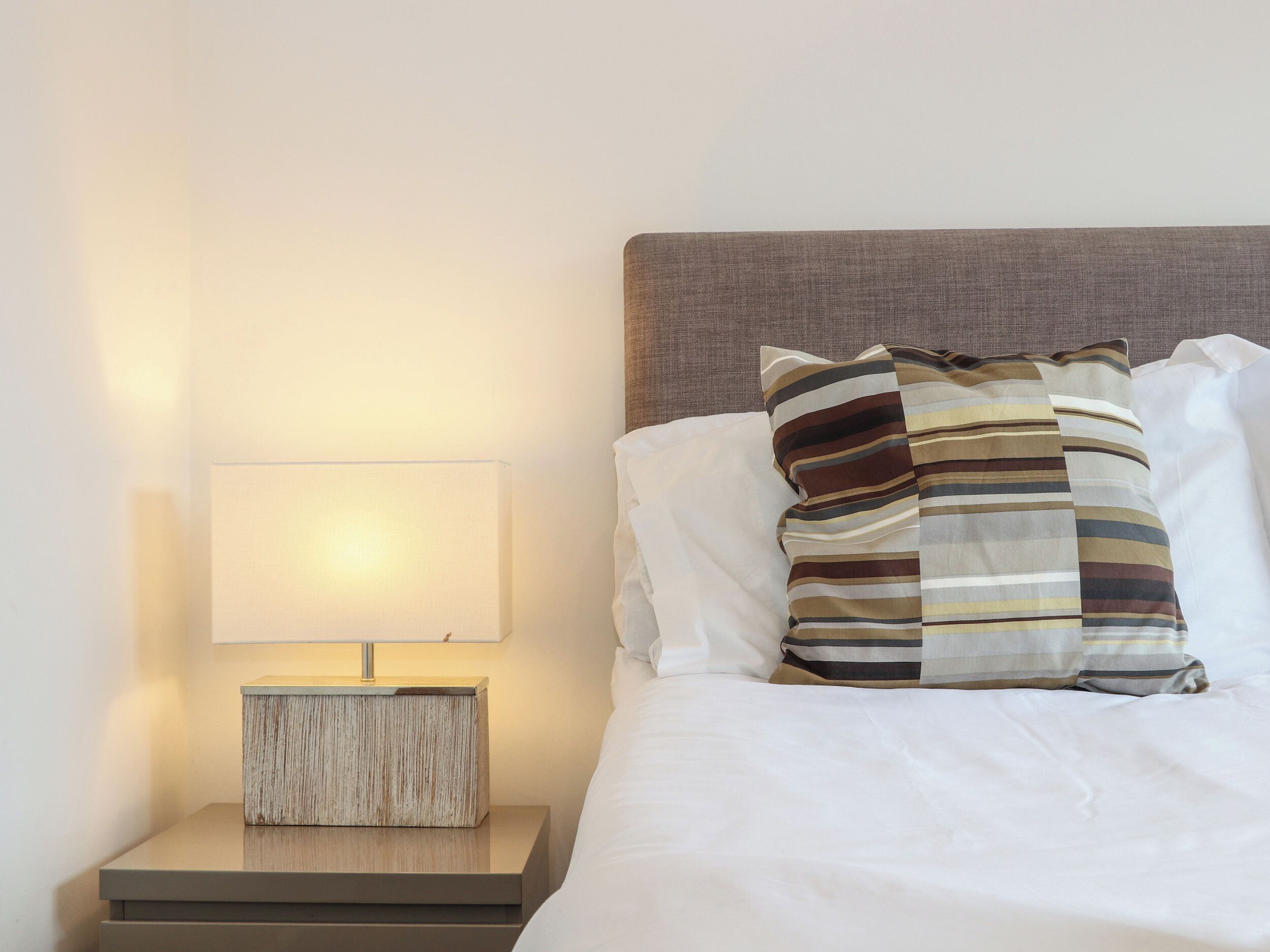 A neatly made bed with white linens and striped pillows, a bedside table with a lamp, and a wall-mounted light.