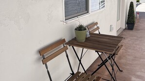 Zona salotto all'aperto con tavolo e panche in legno, una pianta in vaso e una finestra che si affaccia su un edificio in mattoni e vegetazione.