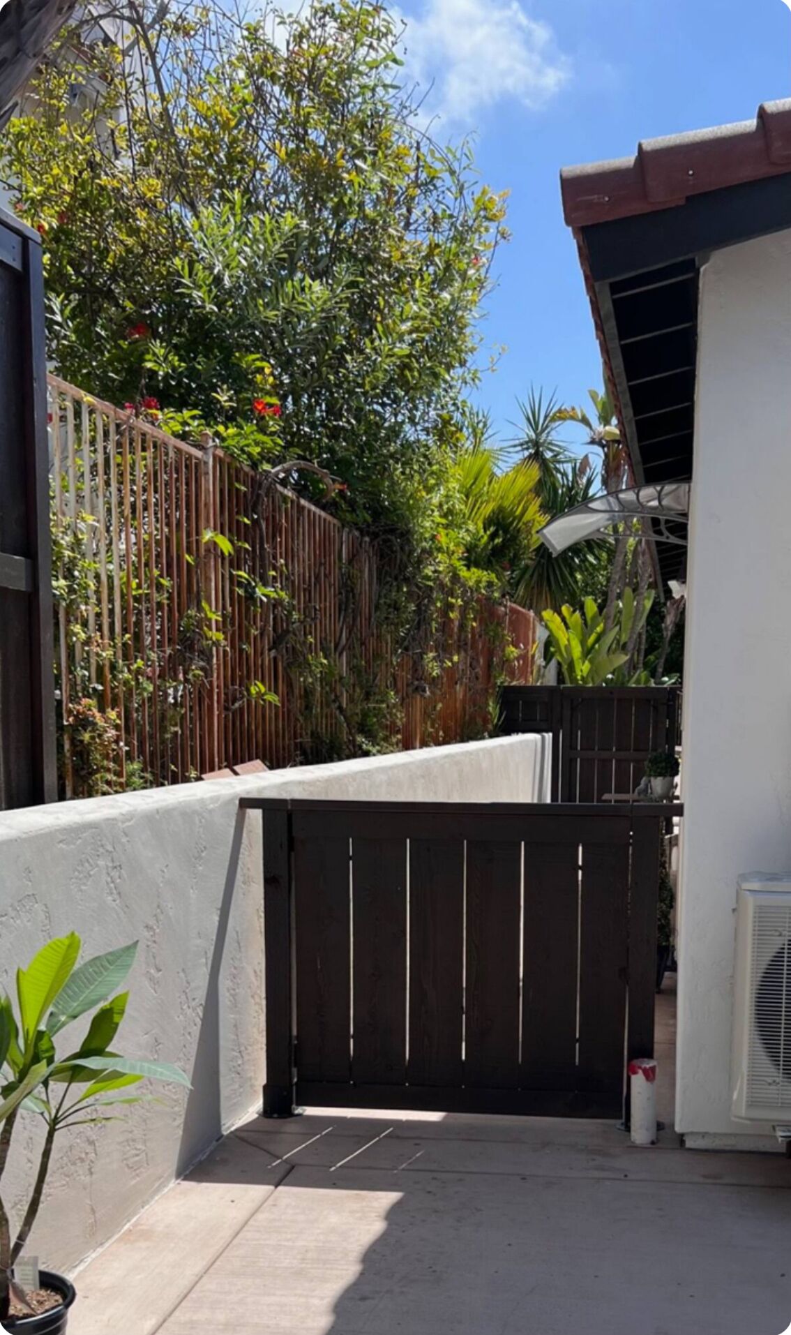Une courClose par un portail, avec une clôture en bois, des plantes en pot et un bâtiment blanc doté d’un toit en pente.