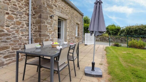 A stone building with a patio table and chairs, a grey umbrella, and a white picket fence. - Emeraude by Interhome (Saint Coulomb)