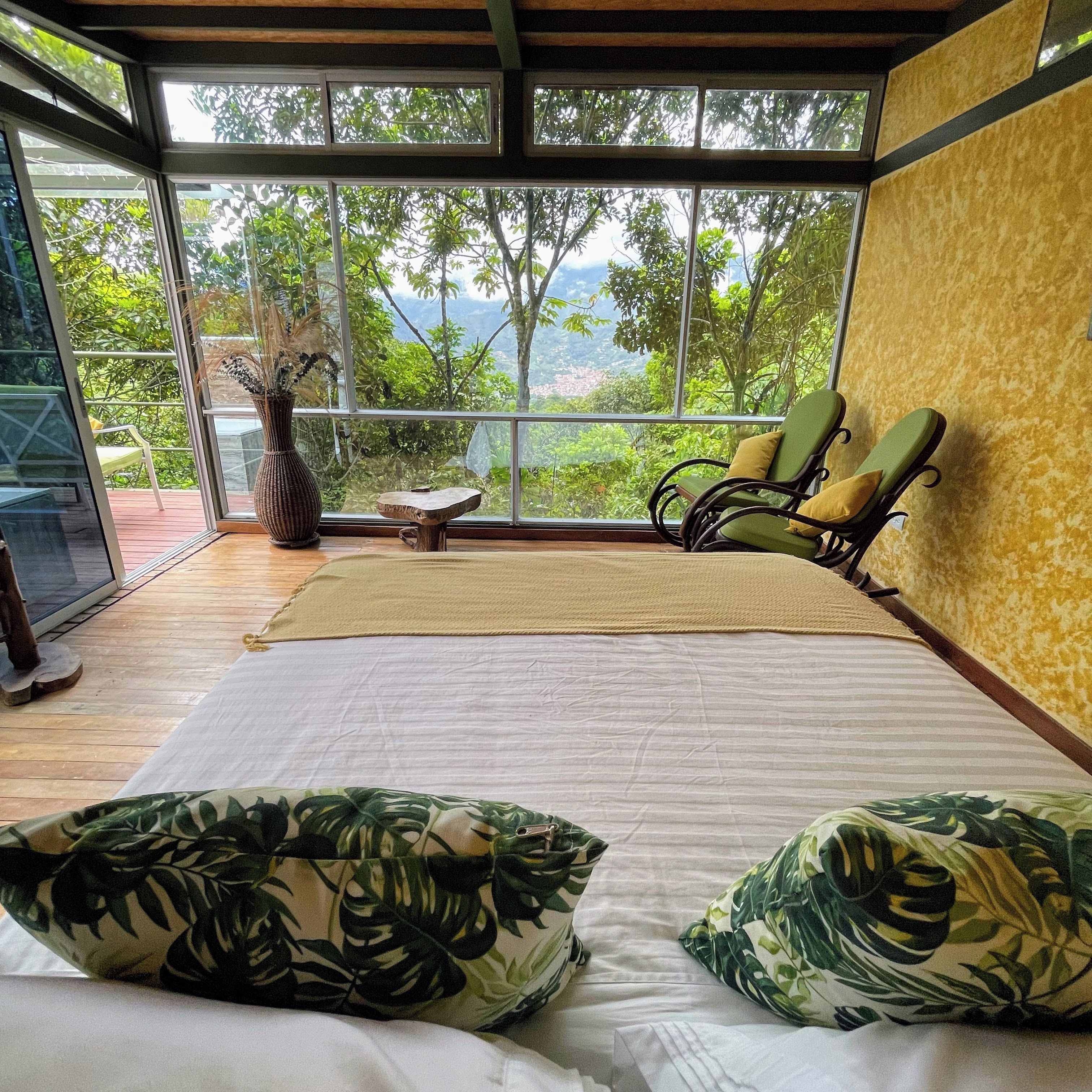 Ein Zimmer mit Bett, Stuhl, Tisch und Blick auf Grün hinter großen Fenstern.