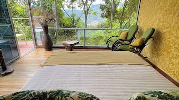 Ein Zimmer mit Bett, Stuhl, Tisch und Blick auf Grün hinter großen Fenstern.