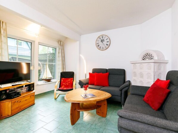 Ein Wohnzimmer mit Fernseher, einem Holztisch, zwei grauen Sofas mit roten Kissen, einem Sessel mit rotem Kissen und einem weißen Wanddekor.