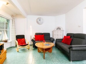 Ein Wohnzimmer mit zwei grauen Sofas, einem Holztisch und einem roten Sessel. Eine große Uhr hängt an der Wand und ein weißes Schrank steht an der Wand.