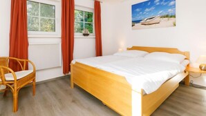 A bedroom with a wooden bed, a wicker chair, a window with curtains, and a beach scene painting on the wall. - Im Kiefernweg DHH 21 by Interhome (Zinnowitz)