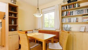 A cozy dining area with a wooden table and wicker chairs, a bookshelf with books and decorative items, and a window with curtains. - Im Kiefernweg DHH 21 by Interhome (Zinnowitz)