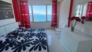 A bedroom with a bed, a bedside table, a chair, a dresser, a window with a view of the sea, and red curtains. - Appartamento in centro con vista e garage (Sarandë)