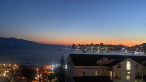 Un paysage urbain côtier au crépuscule, avec des bâtiments le long du front de mer et des bateaux sur l’eau.