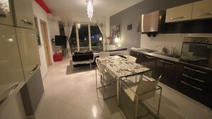A modern kitchen with stainless steel appliances, a dining area with a glass table, and a view of the city at night. - Appartamento in centro con vista e garage (Sarandë)