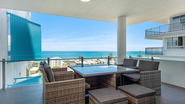 A balcony with a glass table and wicker chairs, overlooking a coastal view.