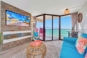 A modern living room with a large TV, a wicker ottoman, a blue sofa, and a view of the ocean through a sliding glass door. - Jacuzzi Corner Wrap Around Balcony Condo Fireplace (Myrtle Beach)