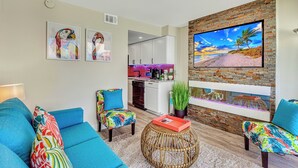 Ein Wohnzimmer mit Sofa, Sesseln, Couchtisch und Kamin. Im Hintergrund ist eine Küche mit weißen Schränken und einer roten Spritzschutzleiste zu sehen.