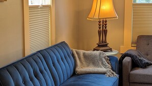 A living room with a blue sofa, a wooden side table with a lamp, a small round rug, and a bookshelf with books. - Da Princess Inn - Walk to Downtown (Lafayette)
