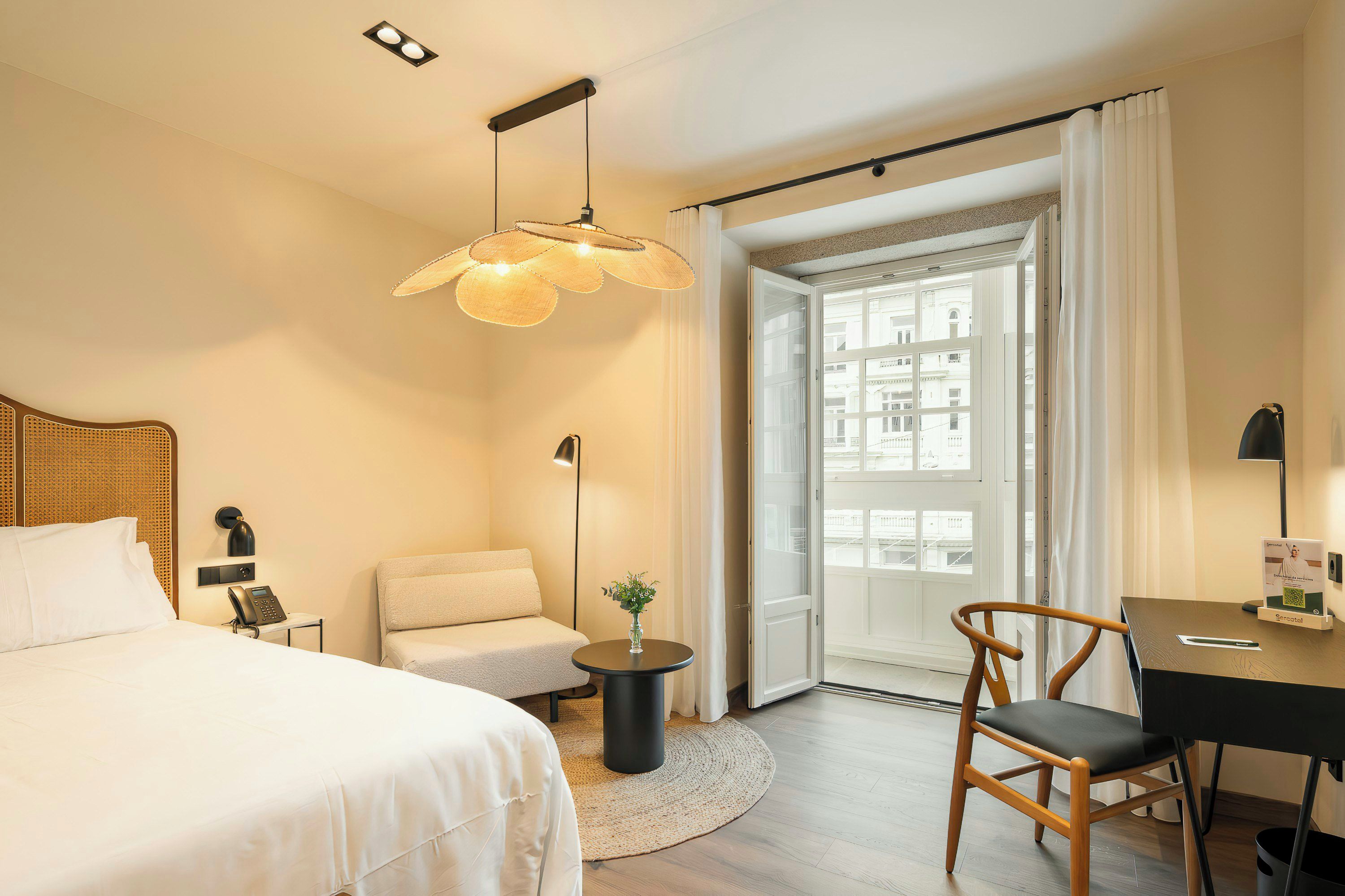 Quarto de hotel com cama, escrivaninha, cadeira, mesinha com um vaso, luminária pendente e vista pela janela com cortinas.