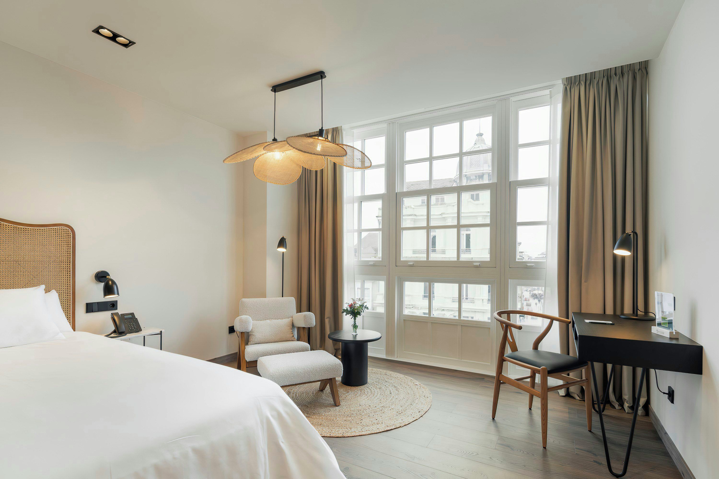 A hotel room with a bed, two chairs, a desk, a small table, and a large window with curtains.