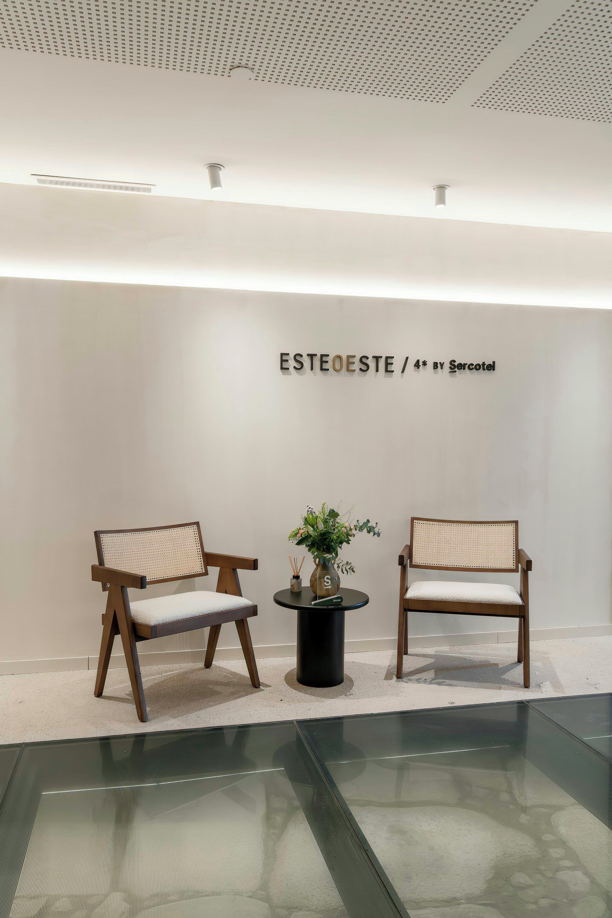 Un intérieur minimaliste avec deux chaises en bois, une petite table surmontée d’un vase et un mur portant l’inscription « ESTEOSTE / 4* BY Sercotel ».