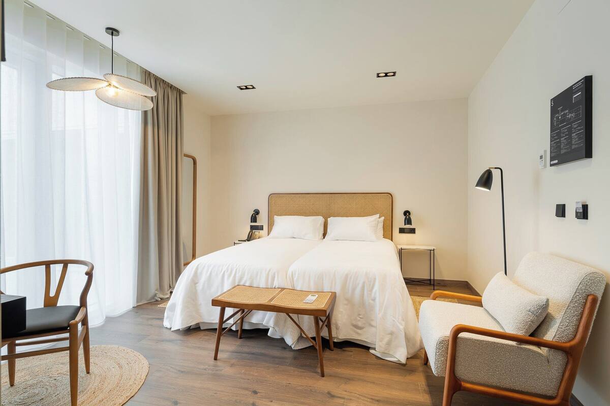 A hotel room with two beds, a wooden chair, a small table, a floor lamp, and a large window with curtains.