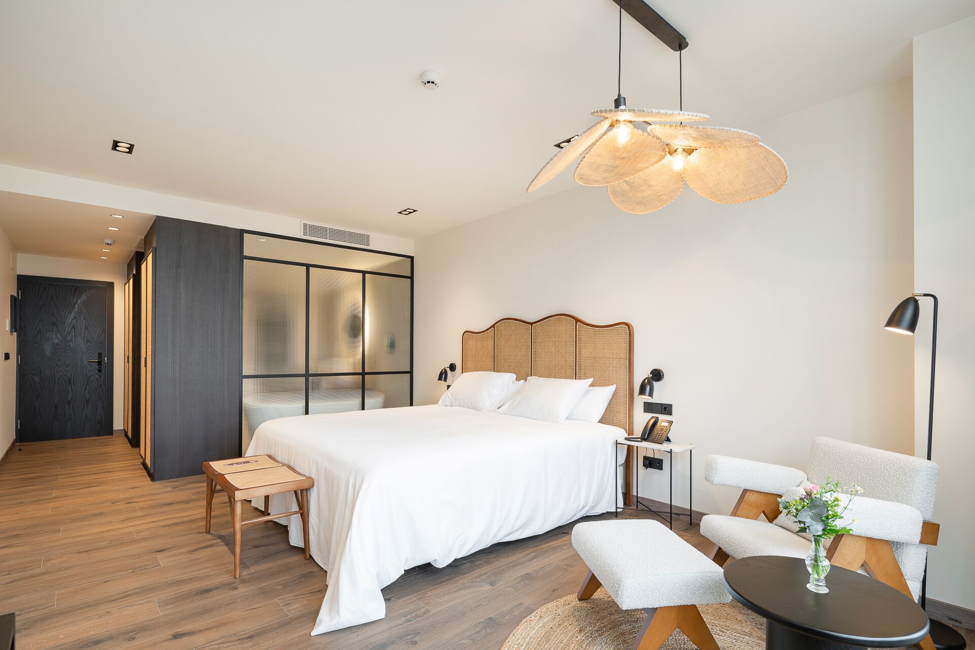 Un dormitorio moderno con una cama grande, un cabecero de madera, una mesita de noche, una silla, una lámpara de pie y un jarrón decorativo.