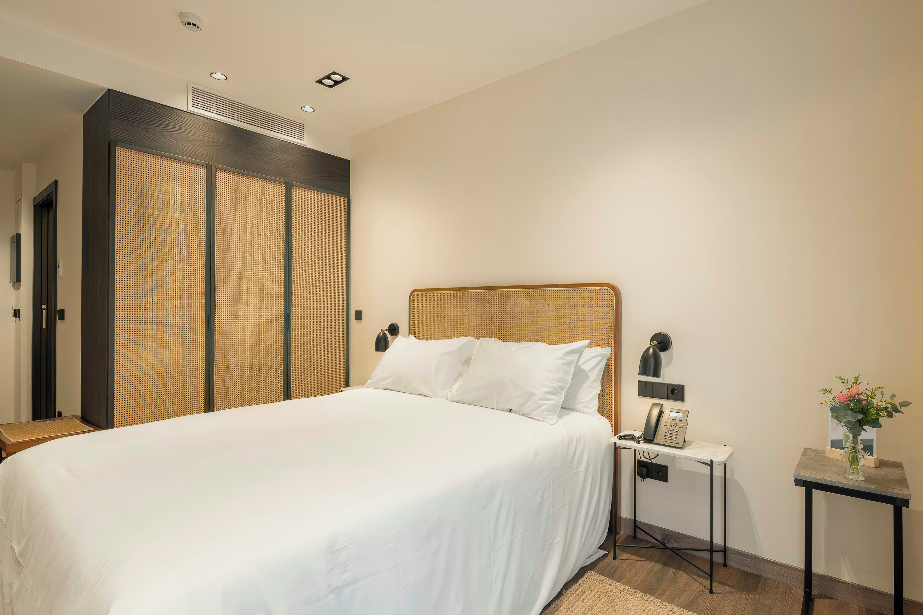 A hotel room with a bed, a bedside table with a phone, a small table with a vase of flowers, and a wall-mounted lamp.