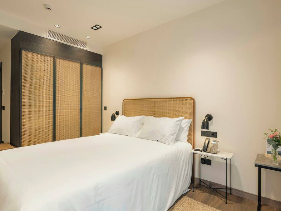 A hotel room with a bed, a bedside table with a phone, a small table with a vase of flowers, and a wall-mounted lamp.