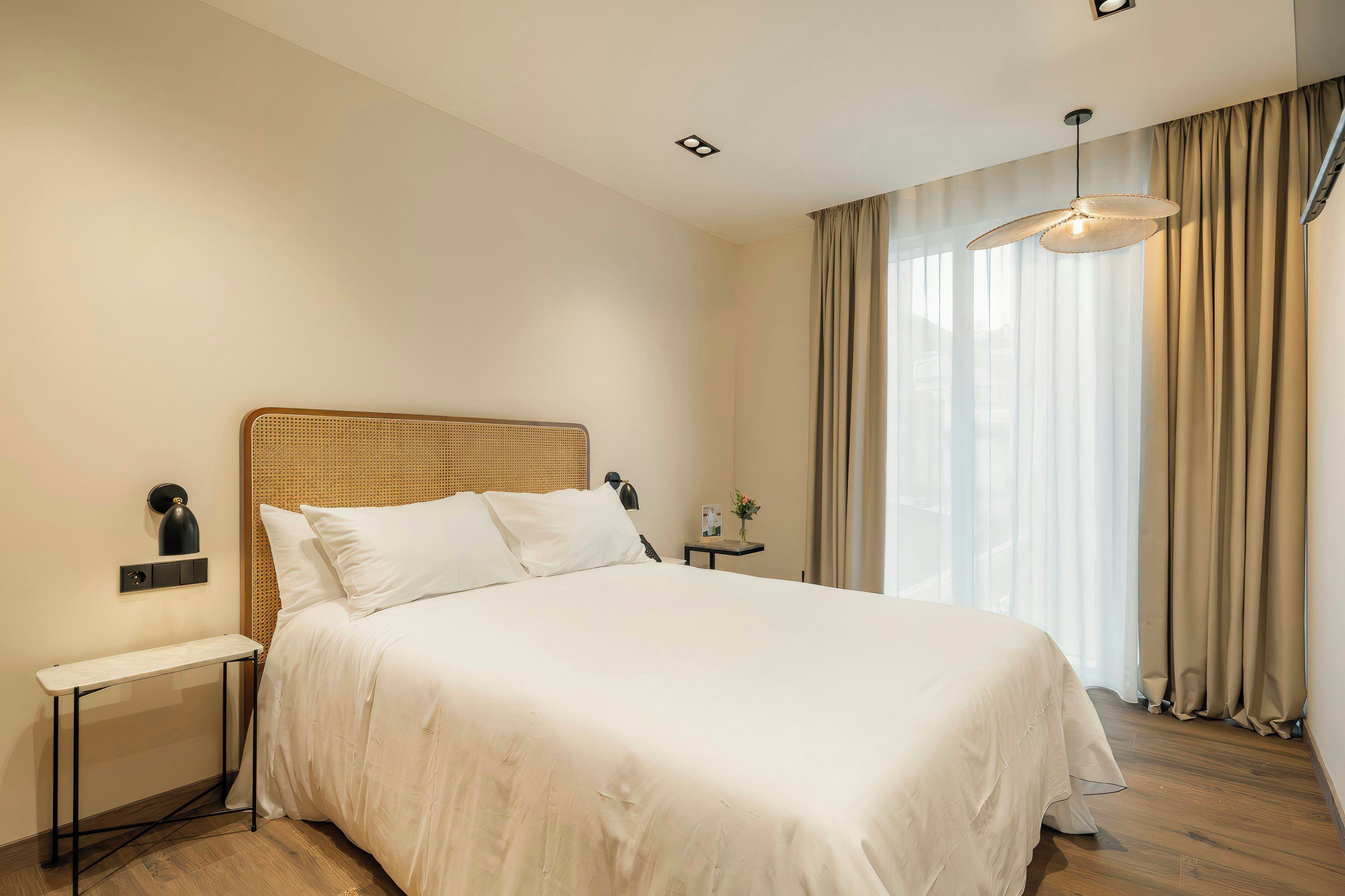 Una habitación de hotel con una cama grande, una mesita de noche, una lámpara, una pequeña mesa con un florero y una ventana con cortinas.
