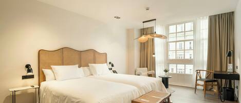 A hotel room with two beds, a desk, a chair, a lamp, and a large window with curtains.