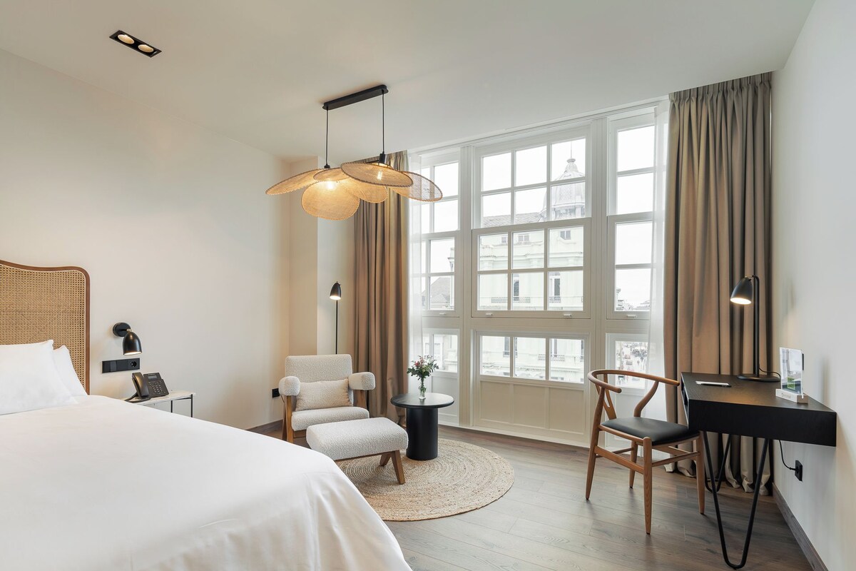 A hotel room with a bed, a desk, a chair, a round rug, a hanging light fixture, and a large window with curtains.