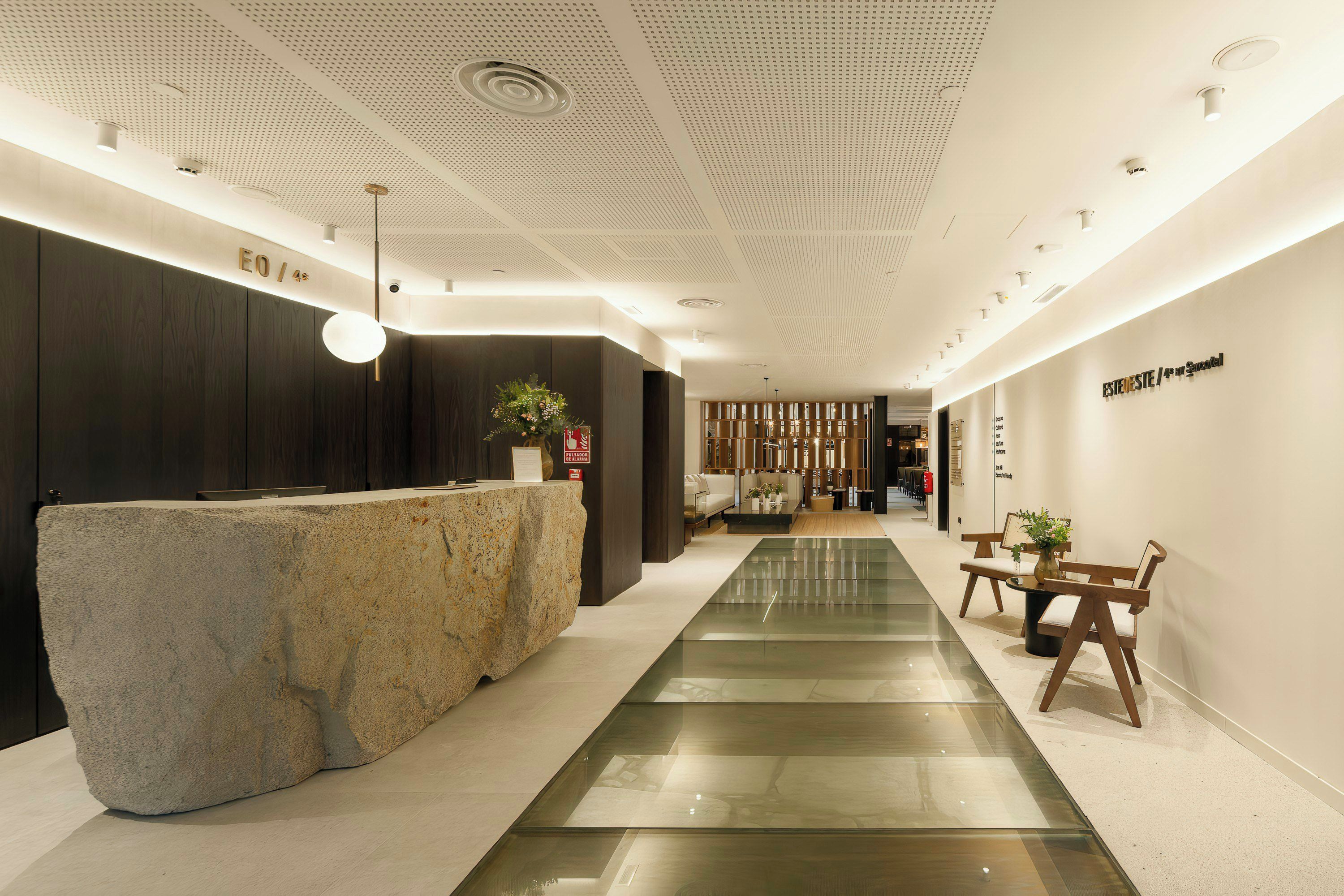 A modern reception area with a stone reception desk, a glass walkway, and a seating area with wooden chairs and tables.
